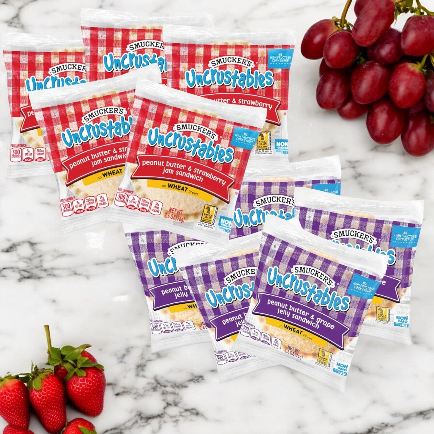 A 10-pack bundle of Uncrustable Smuckers sandwiches, featuring five peanut butter and strawberry jam sandwiches and five peanut butter and grape jelly sandwiches, is displayed on a marble surface next to fresh grapes and strawberries.