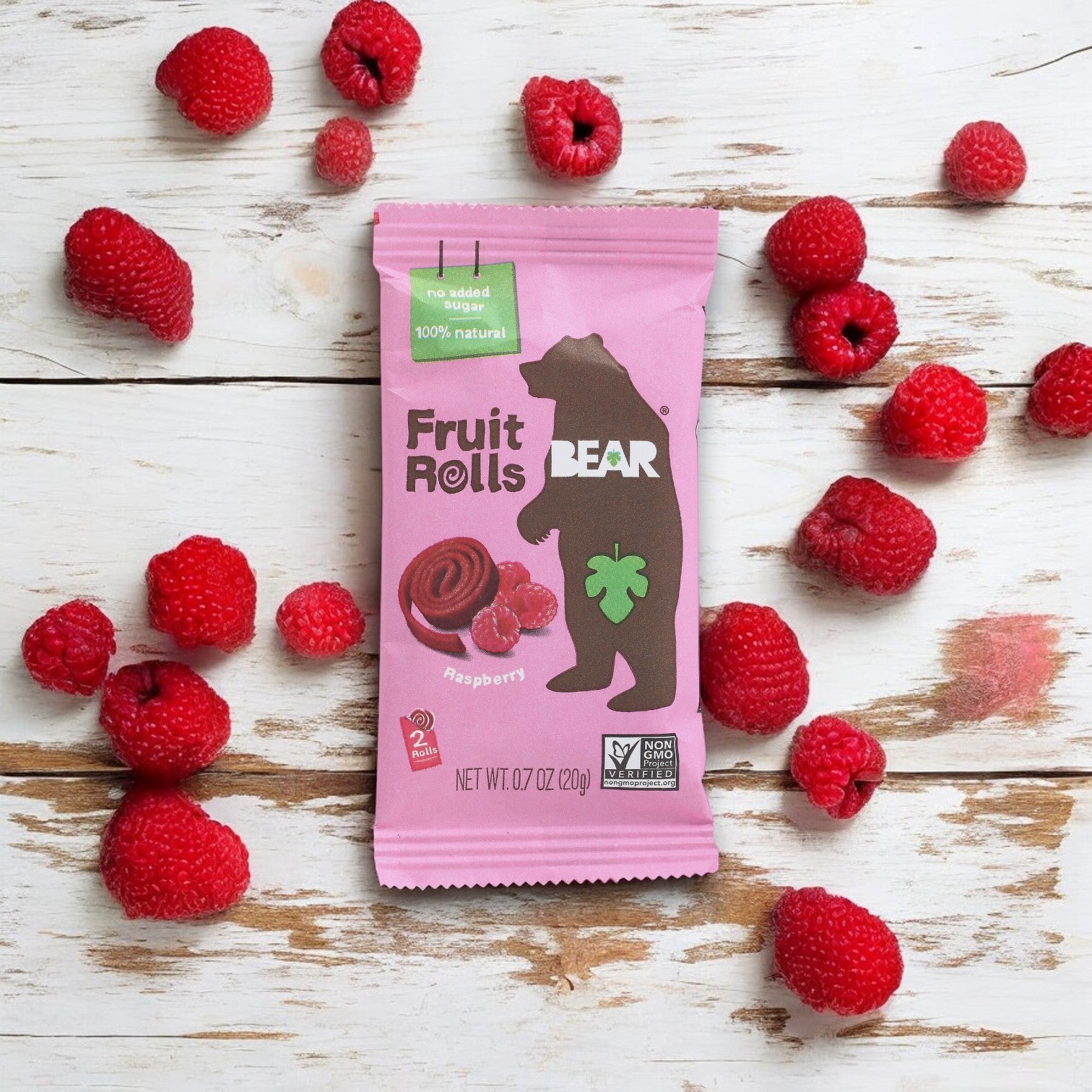 A package of Bear Fruit Rolls - Raspberries, 0.7 oz- 1 Count, sits on a wooden surface surrounded by fresh raspberries. This delicious and healthy treat from Bear is a 100% natural fruit snack with no added sugar.