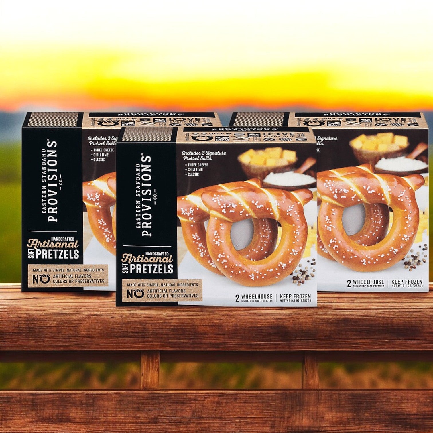 Three packs of Easy Lunches Eastern Standard Provisions Signature Soft Artisanal Handcrafted Pretzels sit on a wooden surface, with a blurred outdoor sunset in the background.