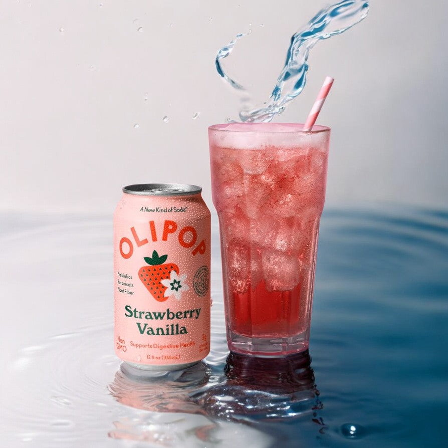 A can of Olipop OLIPOP Prebiotic Soda, Strawberry Vanilla- 12 fl oz - 1 Count stands next to a glass filled with the beverage, ice, and a straw on a reflective surface with a splash of water in the background. This refreshing drink not only delights your taste buds but also supports digestive health with prebiotics.