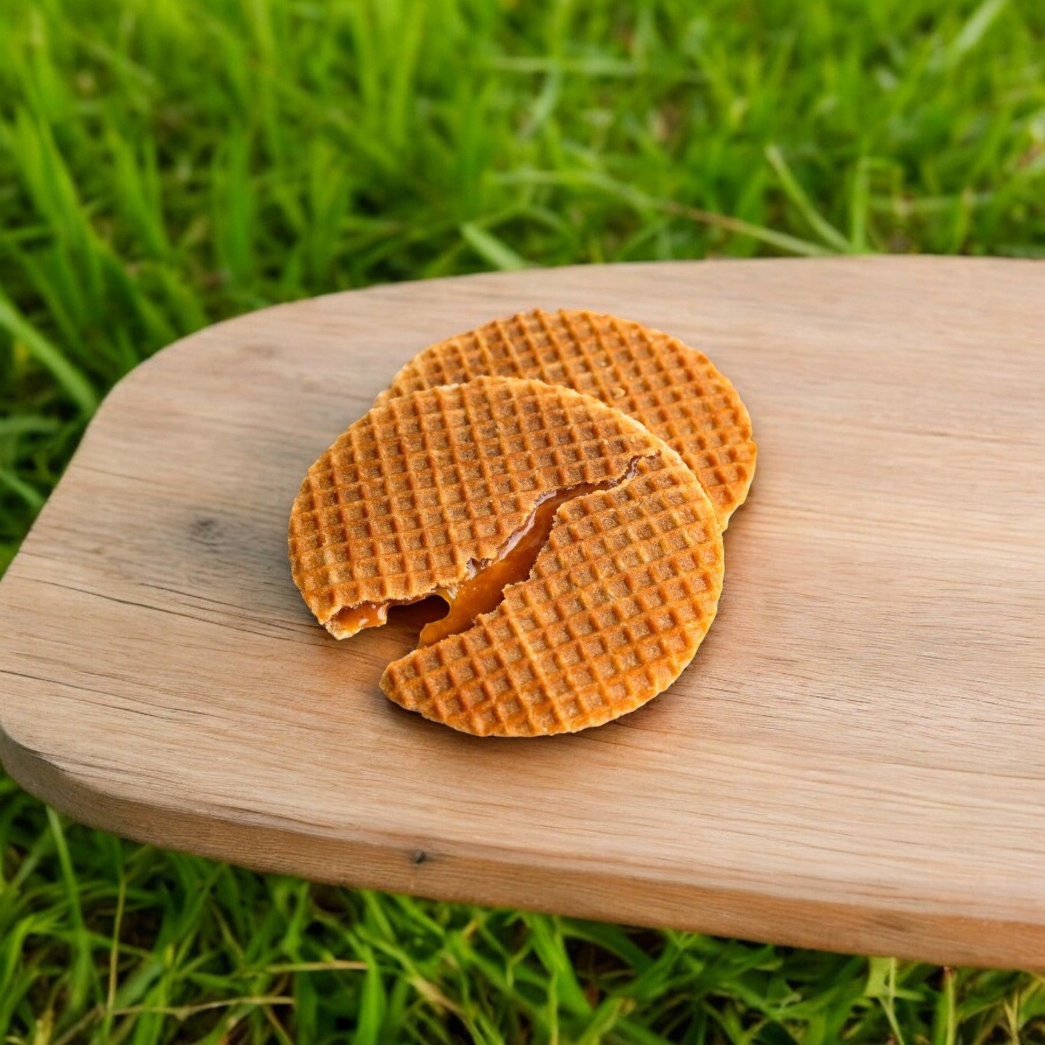 Two Daelmans Individually Wrapped Caramel Stroopwafels (2.75 oz each), one partially cracked, are placed on a wooden board with grass in the background, showcasing this Dutch delight made from natural ingredients.