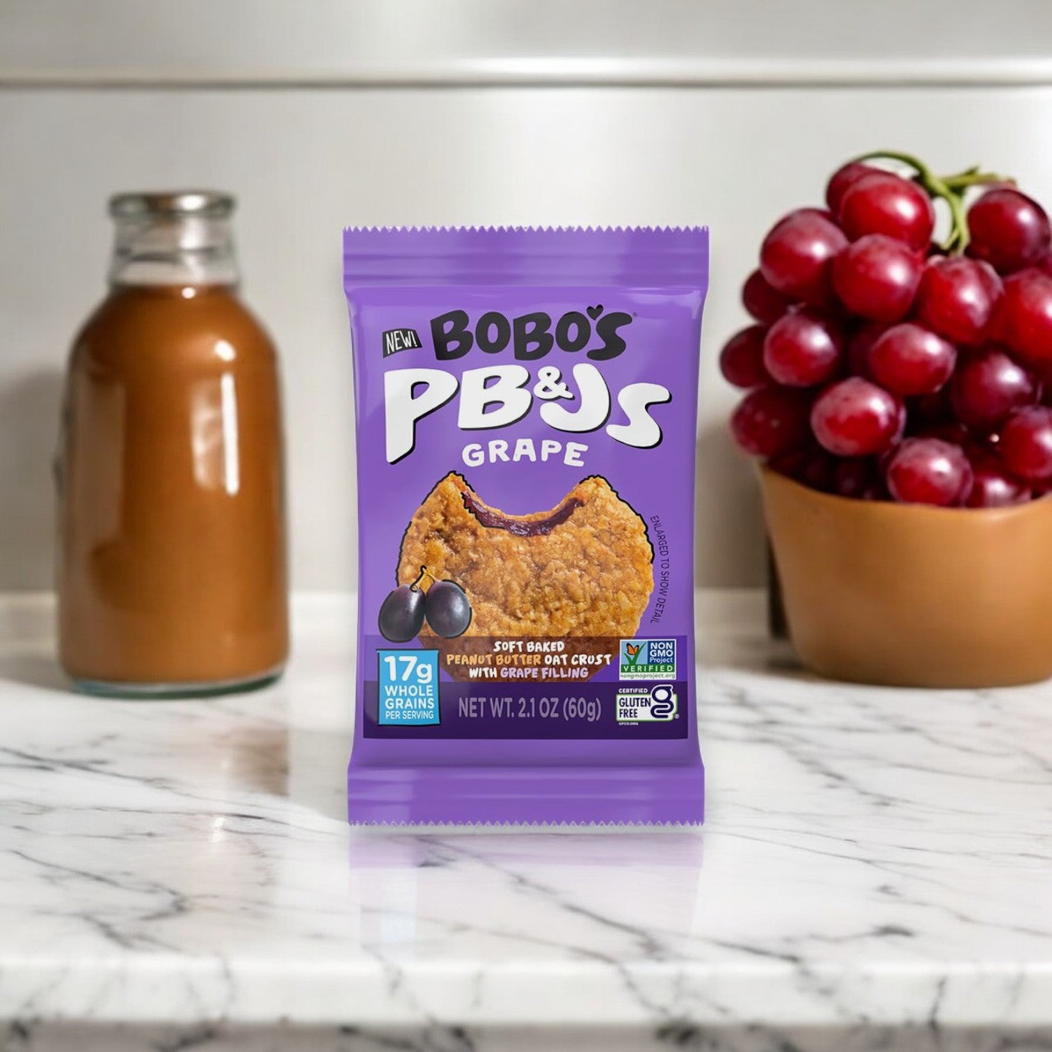 A packet of Bobo's PB&Js Grape, 2.1 oz - 1 Count is placed on a marble countertop next to a small bowl of red grapes and a glass bottle of brown liquid. These gluten-free, portable snacks by Bobo's are perfect for any time you need a quick, delicious pick-me-up.