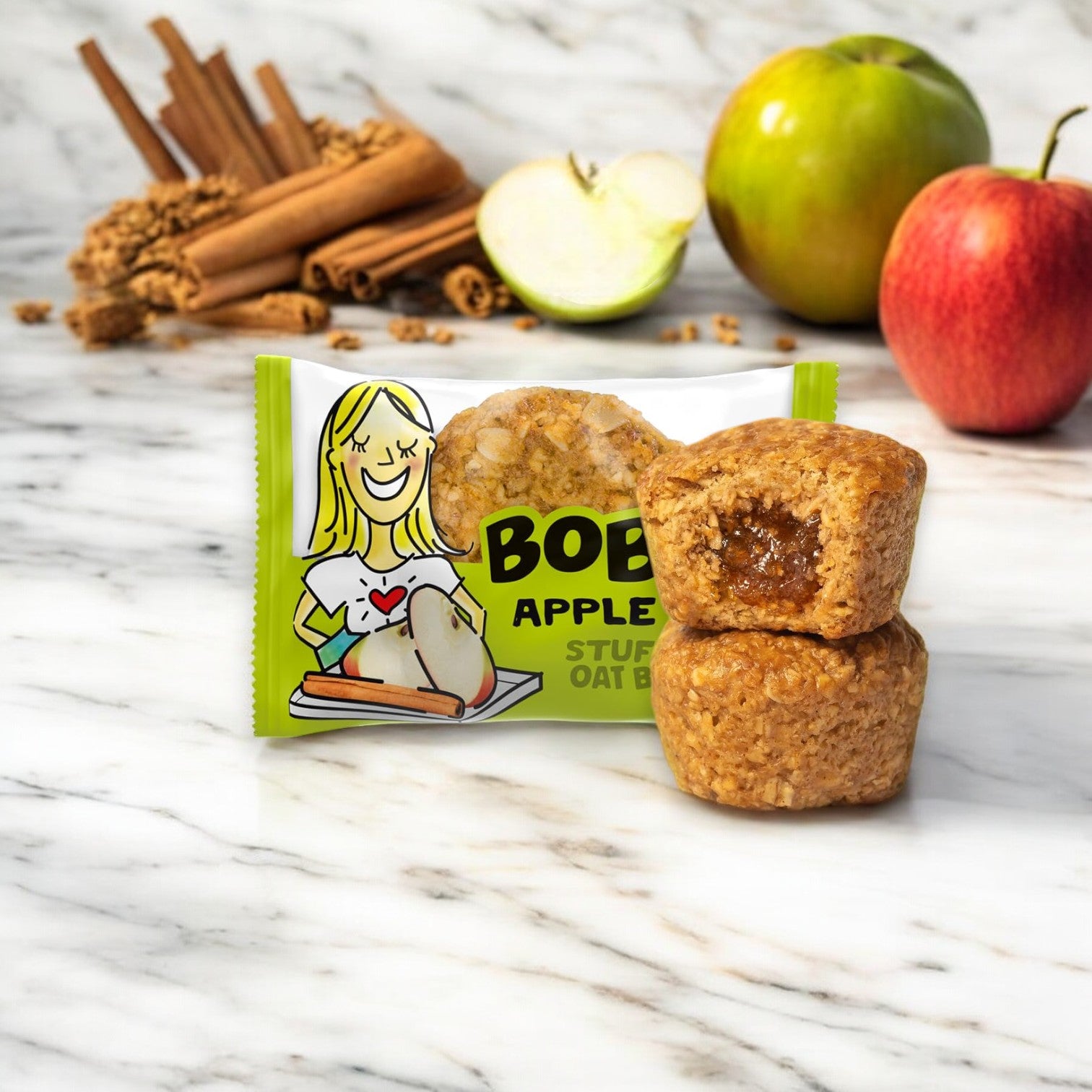 A package of Bobo's Stuff'd Apple Pie Bites is displayed on a marble surface, with two bites in front. Made with wholesome whole grain oats and featuring fresh apples and cinnamon sticks in the background, these bites are both vegan and gluten-free.