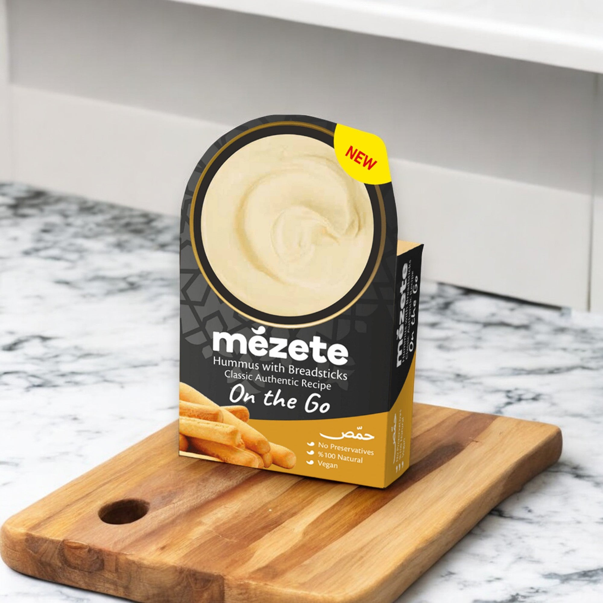 A 3.2 oz package of Mezete Dip and Go Hummus with Bread Sticks sits on a wooden cutting board. Ideal as an on-the-go snack, the packaging highlights its "Classic Authentic Recipe" and "New" status while proudly displaying labels for "No Preservatives," "100% Natural," and "Vegan.