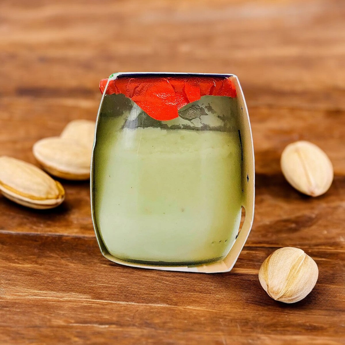 A jar of Pistachio Organic French Dessert by Petit Pot, adorned with a red topping, is placed on a wooden table surrounded by four whole pistachios—an elegant nod to artisanal French cuisine.