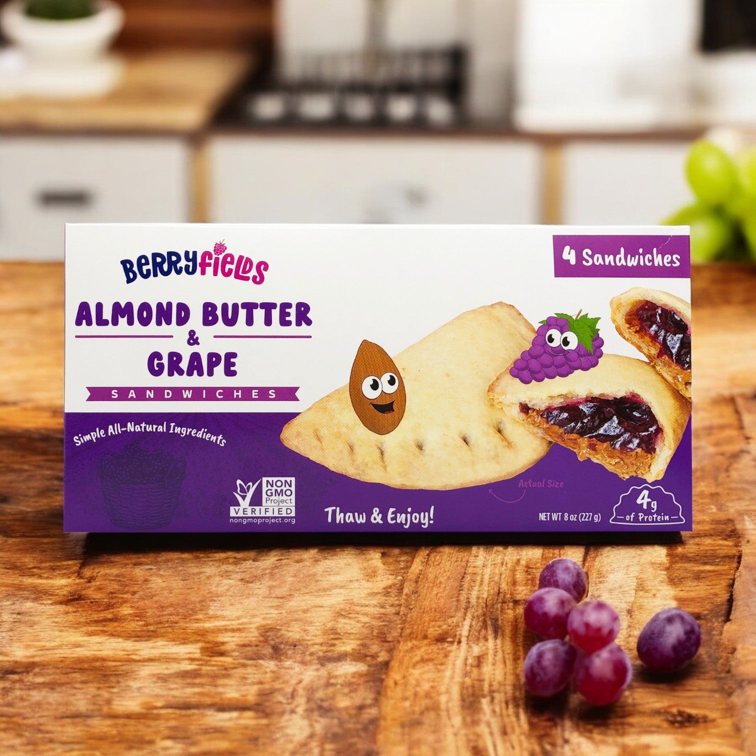 A box of Berryfields Variety Pack, featuring one Almond Butter & Grape Sandwich and one Almond Butter & Strawberry Sandwich, sits on a wooden table with a few grapes beside it.