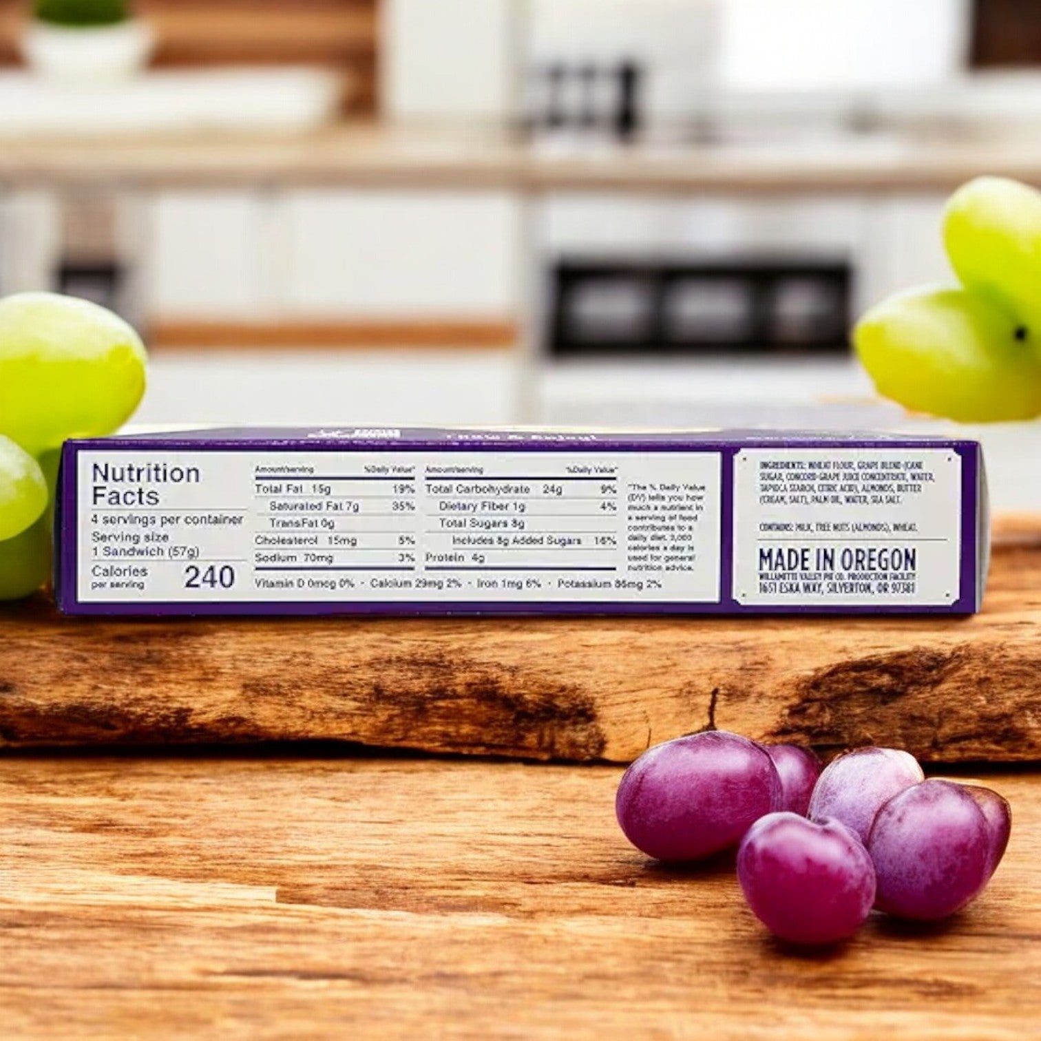 A package of Berryfields Almond Butter & Grape Sandwiches, 8 oz. - 3 Count, placed on a wooden surface with green and purple grapes nearby, complements the rich taste of almond butter.