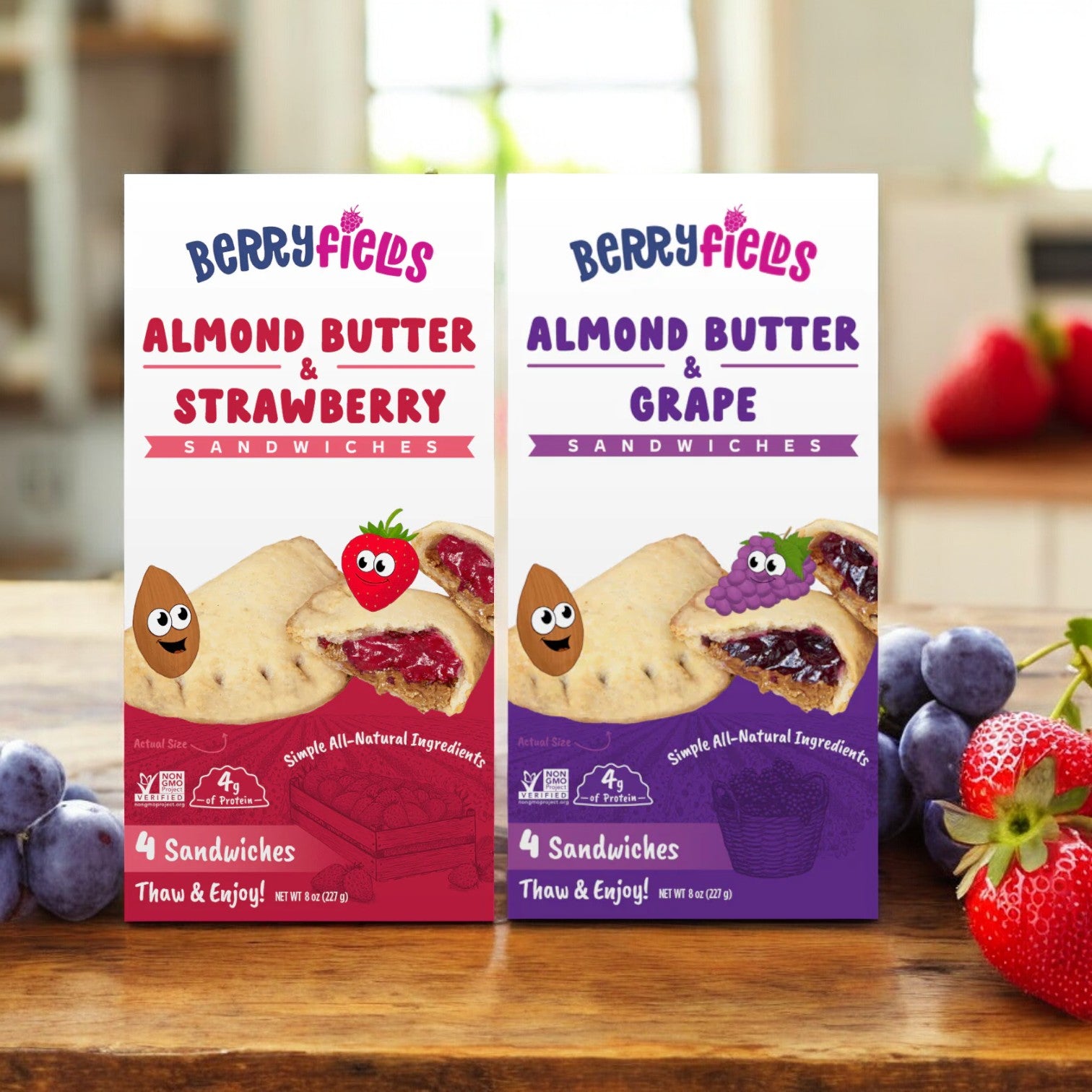 A Berryfields Variety Pack with one box of Almond Butter & Strawberry Sandwiches and another of Almond Butter & Grape Sandwiches is arranged on a wooden table, surrounded by fresh strawberries and grapes.