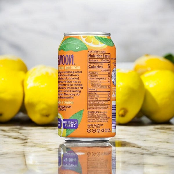 A can of Swoon Half & Half Black Tea and Lemonade, sugar-free and monk fruit sweetened, is placed on a reflective surface with several lemons in the background, showcasing its nutritional information.