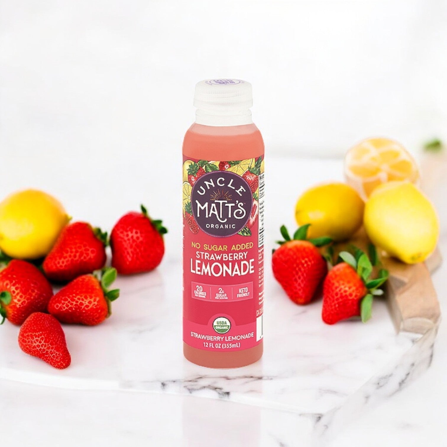 A bottle of Uncle Matt's Organic No Sugar Added Strawberry Lemonade, 12 oz - 1 Count is placed on a marble surface, surrounded by fresh strawberries and whole lemons, highlighting this refreshing beverage from Uncle Matt's sweetened with organic stevia.