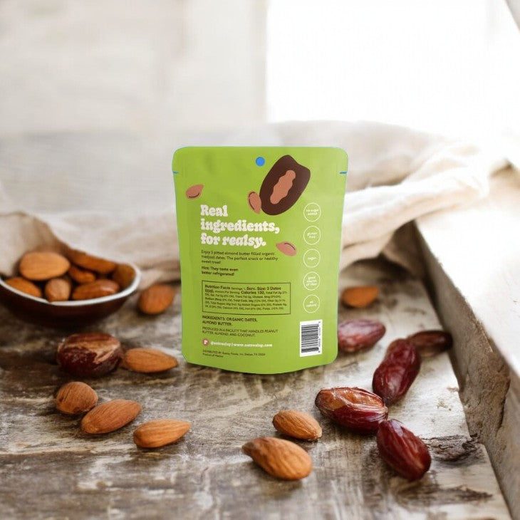 A small green bag of Realsy Almond Butter Dates, crafted from naturally sweet produce sourced from a family-owned date farm, is placed on a wooden surface surrounded by almonds and dates. The bag features the slogan "Real ingredients, for Realsy." along with the nutritional information on the back.