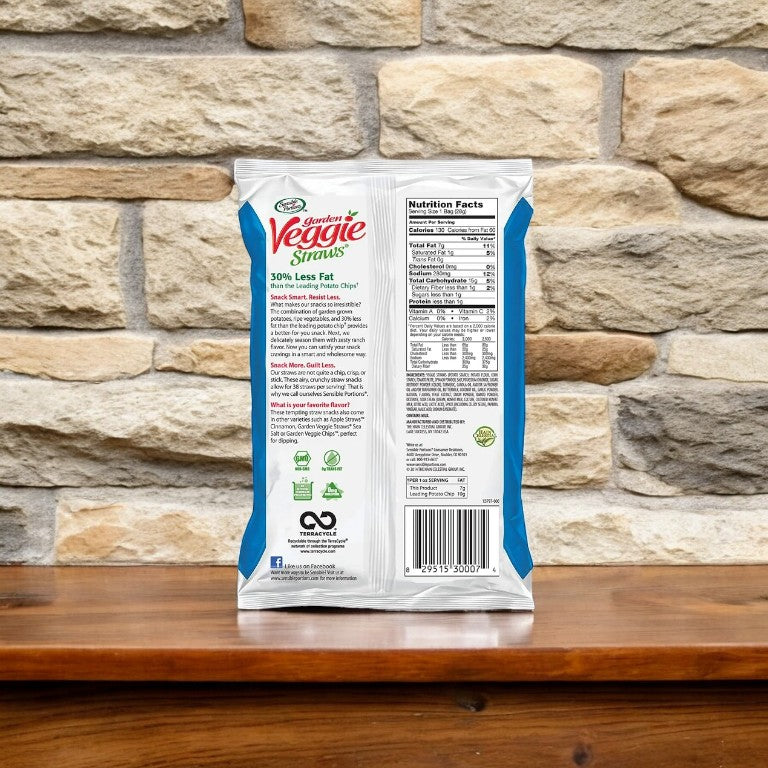 Back view of a Hain Celestial Garden Veggie Straws Gluten-Free Zesty Ranch snack bag showing nutritional facts and ingredients, placed on a wooden surface against a stone wall background. Enjoy these Non-GMO Snacks that are also Gluten-Free, perfect for any health-conscious snacker.