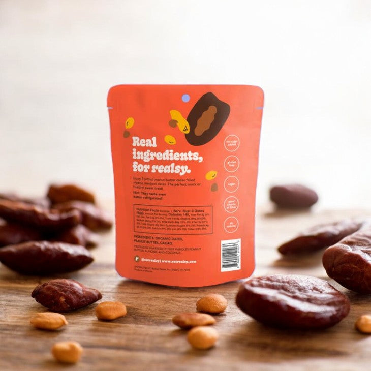 A red packet of Realsy Cacao Peanut Butter Dates with the text "Real ingredients, for realsy" on a wooden surface, surrounded by scattered dates and almonds, showcasing a healthy alternative to Snickers made from naturally sweet produce.