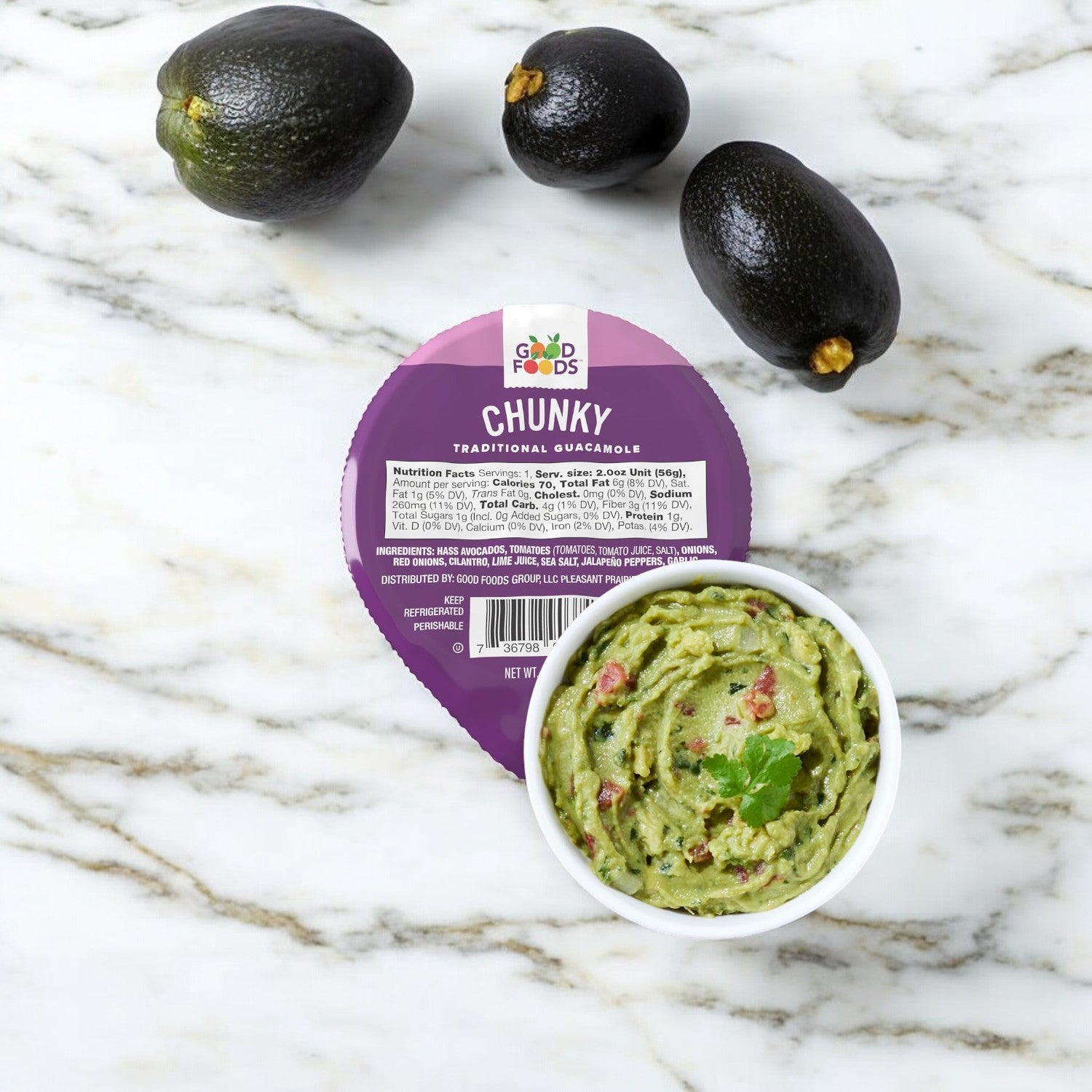 A half-ounce container of Good Foods Chunky Guacamole and a bowl of this gluten-free, healthy snack are on a marble surface, surrounded by three whole avocados.