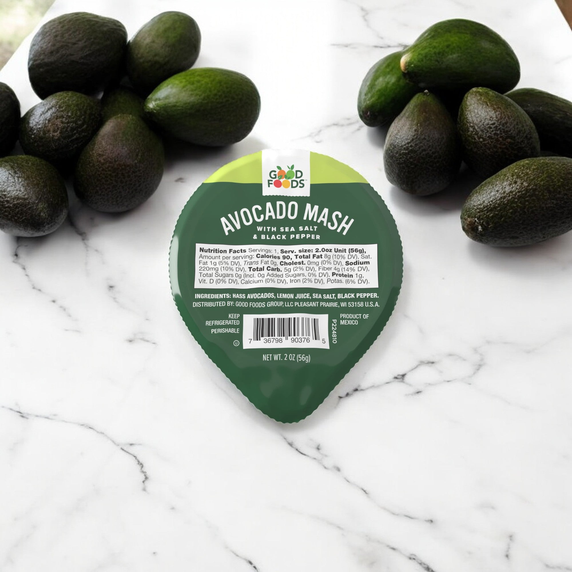 A single-serving container of Good Foods Organic Avocado Mash, Sea Salt & Black Pepper, 2 oz is placed on a white surface, surrounded by several whole avocados.