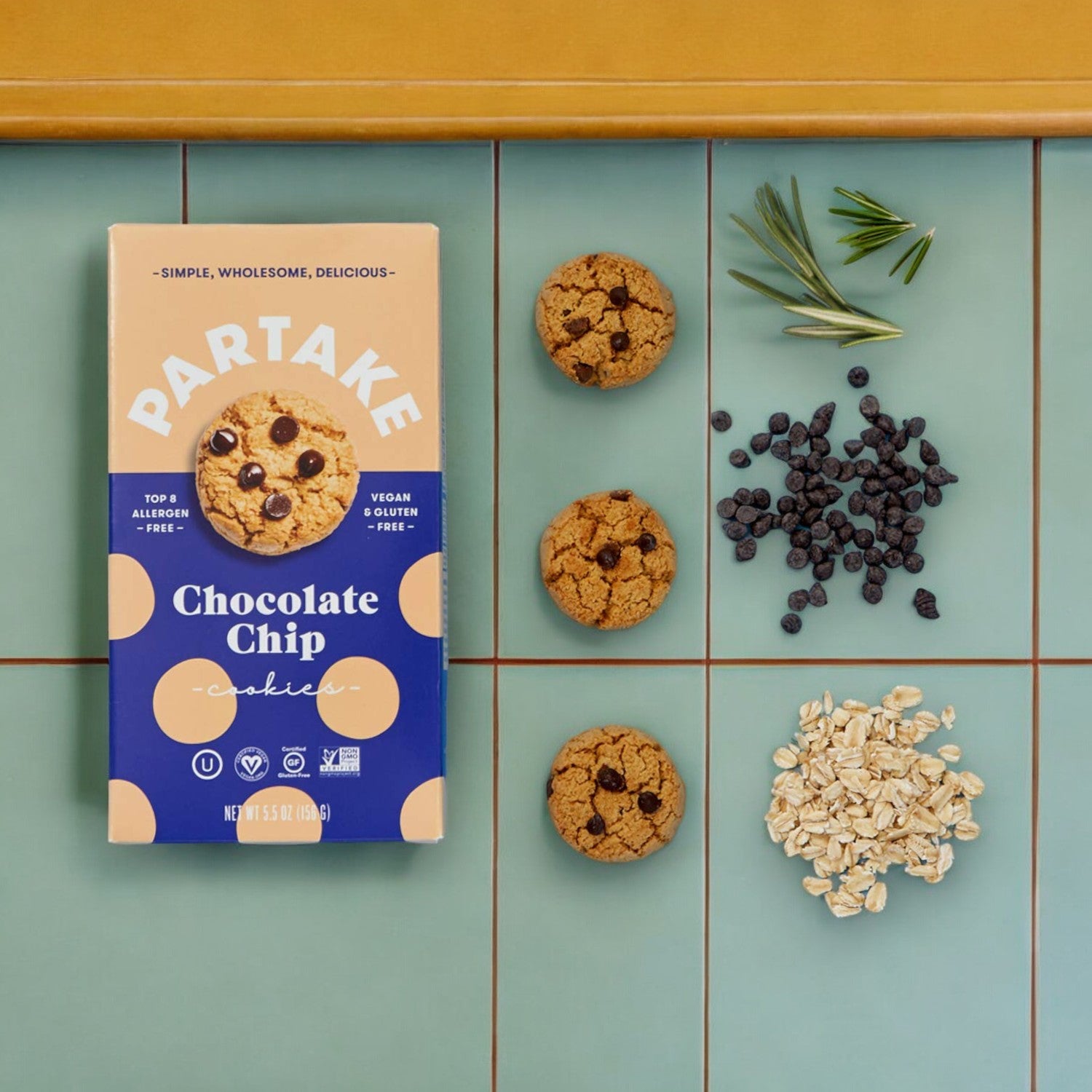 A box of Partake Gluten Free Vegan Chocolate Chip Cookies from the brand Partake is placed on a tiled surface along with three cookies, oats, dried blueberries, and herbs, showcasing our commitment to sustainability and community support.