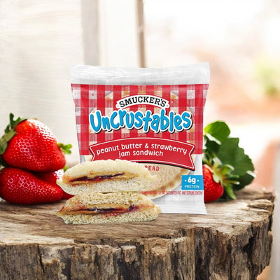 An unopened package of Uncrustable Smuckers' Uncrustables Peanut Butter & Strawberry Jelly, 10 Count - 2 Packs (totaling 20 sandwiches) sits on a wooden surface, with a split open sandwich and fresh strawberries displayed beside it.