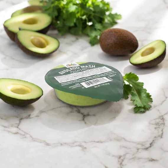 A sealed container labeled "Good Foods Organic Avocado Mash, Sea Salt & Black Pepper, 2 oz" on a marble surface, surrounded by halved avocados and a bundle of cilantro.