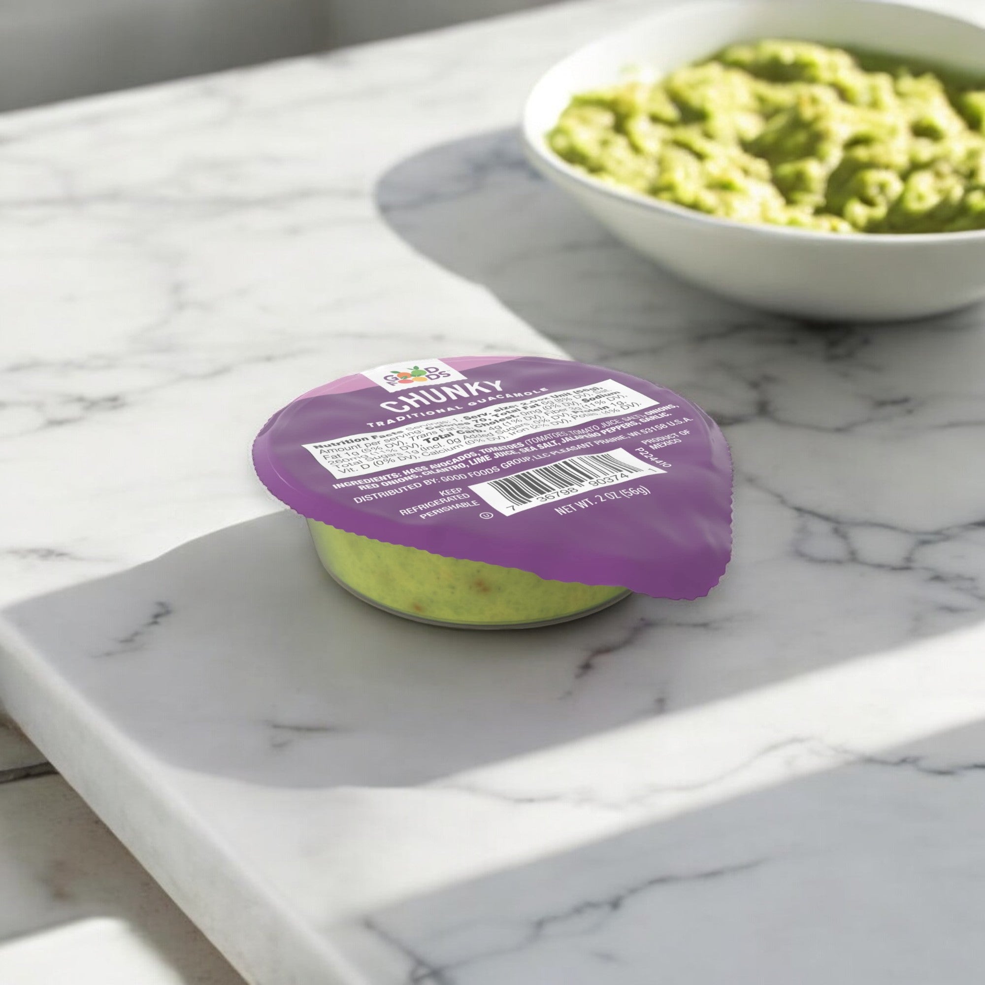 A sealed container of Good Foods Chunky Guacamole - 1/2 oz, featuring a distinct purple lid, sits on a marble counter. A bowl of this healthy, gluten-free snack is in the background, ready to be enjoyed.