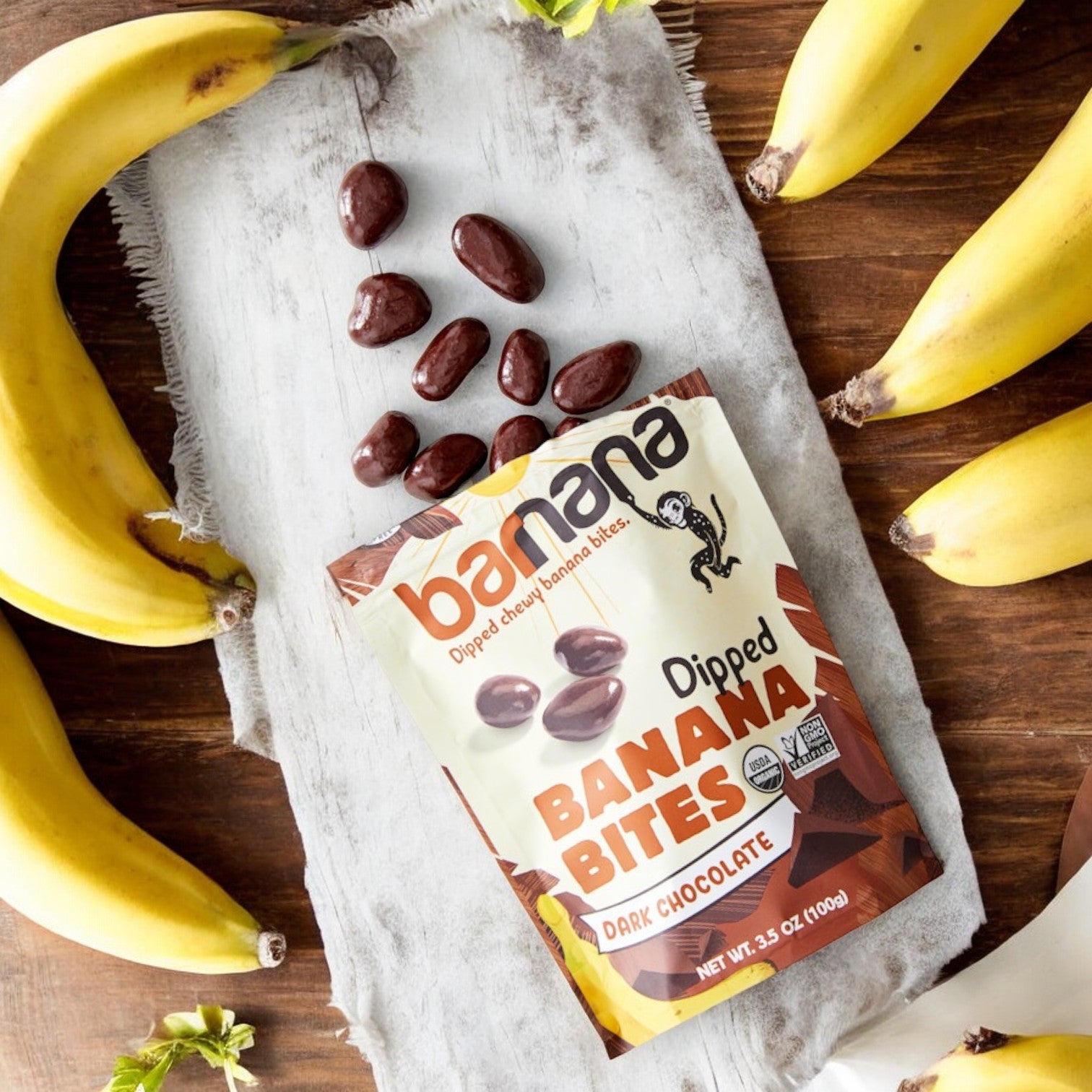 On a wooden surface, surrounded by whole bananas, sits a bag of Barnana Banana Bites Organic Chewy Chocolate. The 3.5 oz bag from Barnara is open, with several delectable, gluten-free snack bites spilled out.