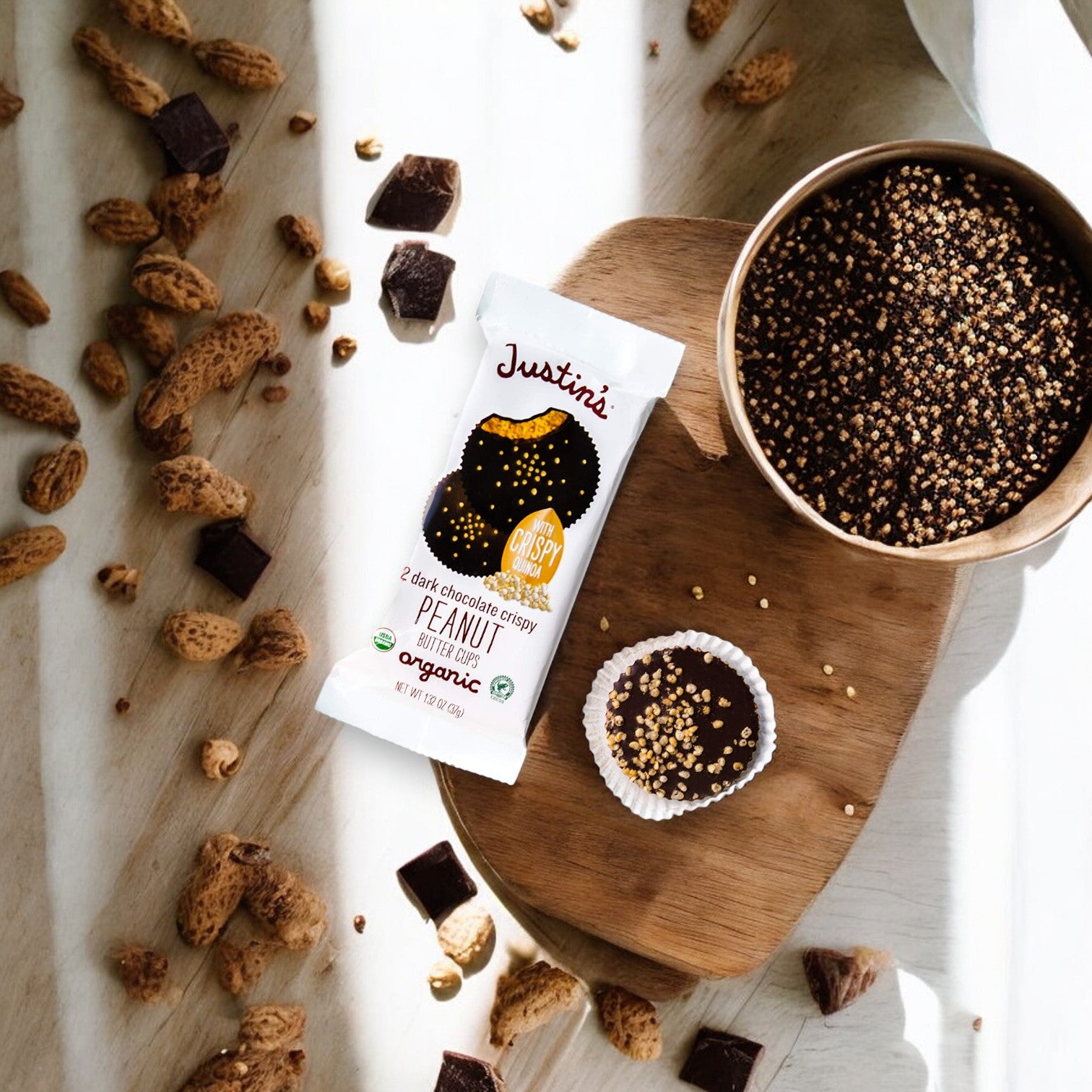 A packet of Justin's Organic Dark Chocolate Peanut Butter Cups-1.4 oz- 1 Count lies on a wooden surface surrounded by peanuts, quinoa, and chocolate chunks.