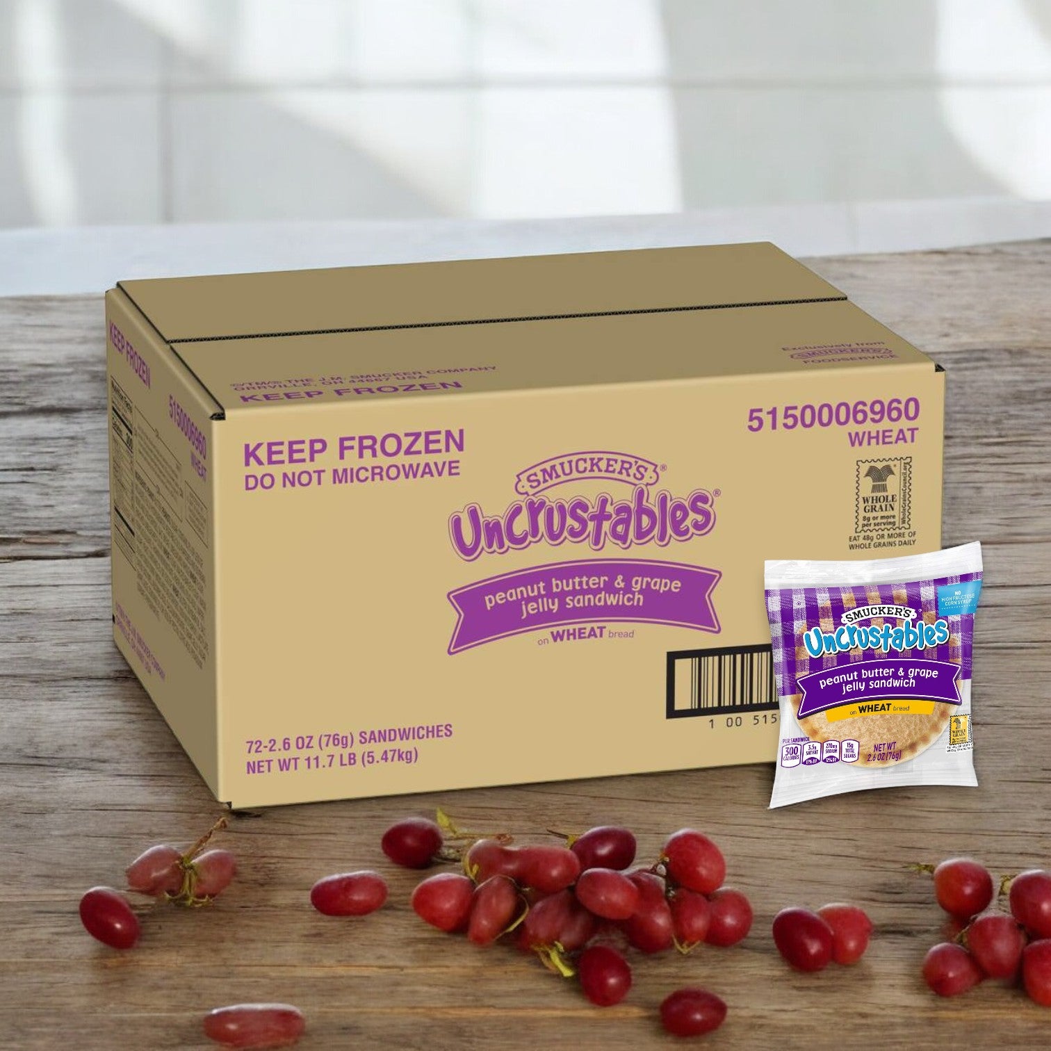 A cardboard box of Uncrustable Smuckers Uncrustables Grape Jam - 72 Count per case sits on a table next to scattered grapes, with a single wrapped sandwich in the foreground, showcasing the perfect blend of creamy peanut butter and sweet grape jam.