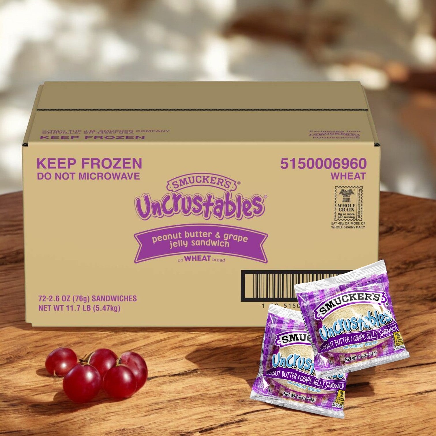 A box of Uncrustable Smuckers Grape Jam sandwiches, containing 72 per case, sits on a wooden surface, with a few loose sandwiches and sweet grapes in the foreground.