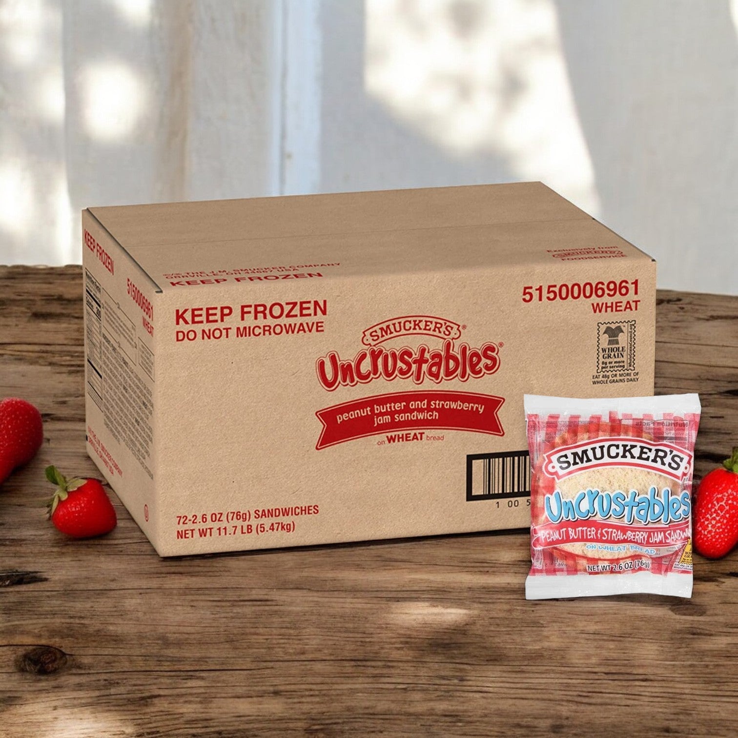 A cardboard box of Uncrustable Smuckers Uncrustables Strawberry Jam - Bulk 72 Count sits next to a single wrapped sandwich and some strawberries on a wooden surface, offering a harmonious blend of flavors. These handheld snacks are perfect for any time of day.