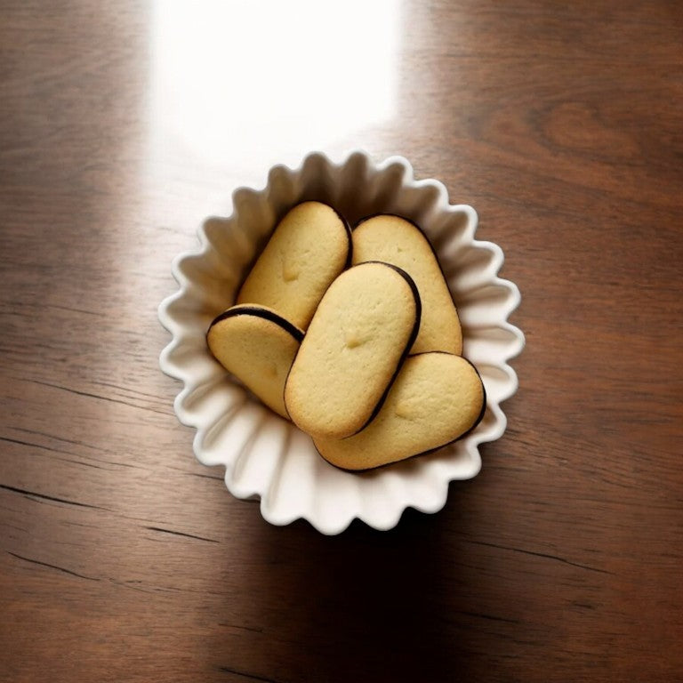 A white ceramic bowl on a wooden table contains several oval-shaped Pepperidge Farm Milano Cookies, Double Dark Chocolate, featuring light brown tops and dark brown edges, made from quality ingredients.