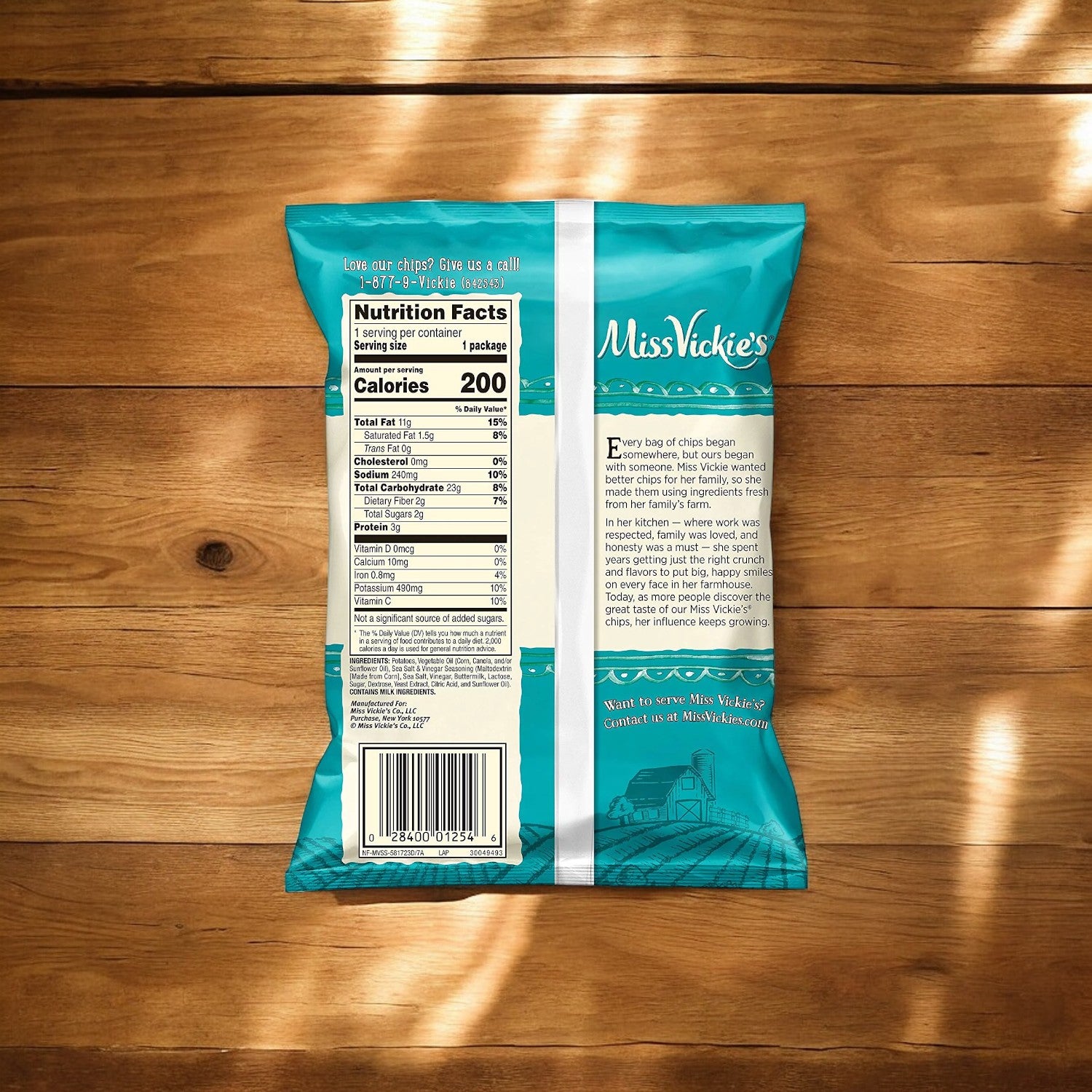 A teal bag of Miss Vickie's Sea Salt & Vinegar Flavored Kettle Cooked Potato Chips (1.375 oz) is displayed with the nutrition facts label facing outward. The bag is positioned on a wooden surface that exhibits patterns of light and shadow, suggesting a natural snacking experience infused with tangy vinegar seasoning.