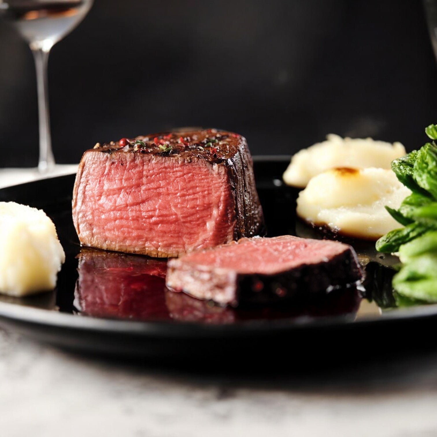 An 8-9 oz Mino Prime Steaks filet mignon, cooked medium-rare, is served on a black plate with creamy mashed potatoes and vibrant greens, while wine glasses are elegantly placed in the background.