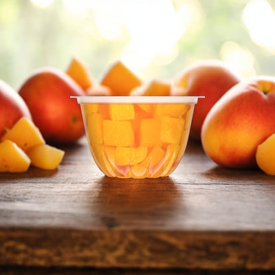 A plastic container of Dole Fruit Bowls Diced Peaches in 100% Fruit Juice, 4 oz (4 Cups), rests on a wooden surface. Whole peaches and peach slices are arranged around the container. The background is softly blurred with natural light, accentuating the freshness preserved by the 100% fruit juice in these delicious fruit bowls from Dole.