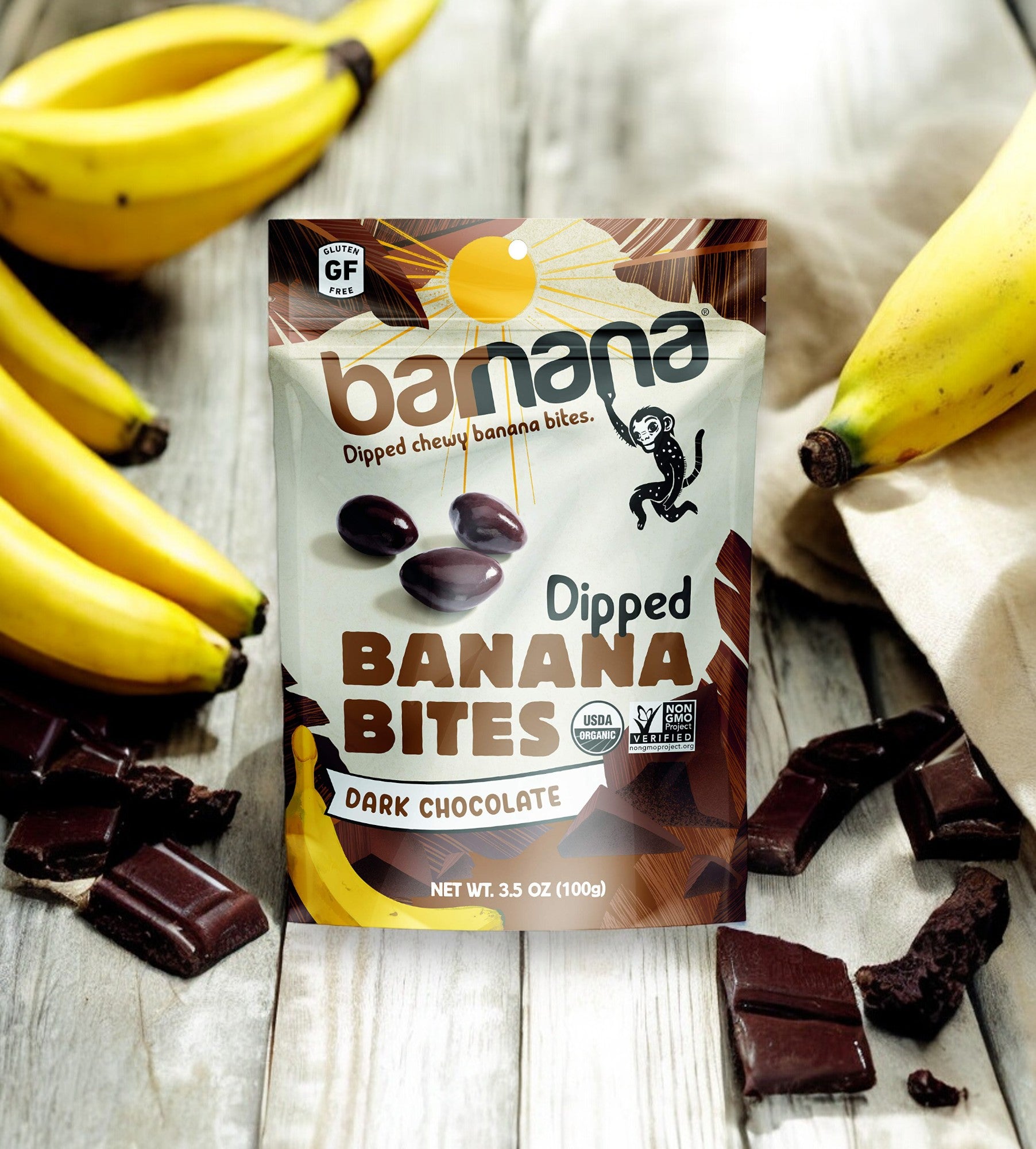 A bag of Barnana's Organic Chewy Chocolate Banana Bites, 3.5 oz, rests on a wooden surface surrounded by fresh bananas, a cloth, and scattered pieces of dark chocolate—creating the ideal sustainable snack.