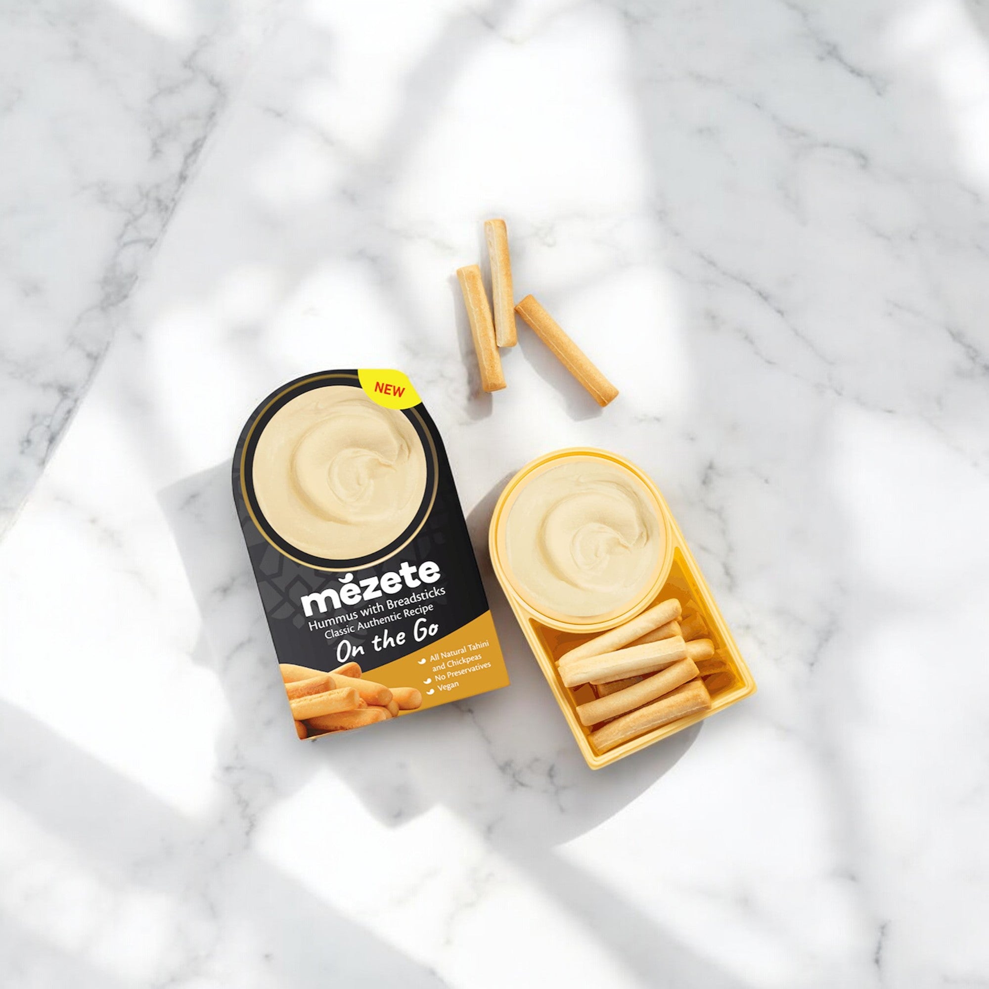 A container of Mezete Dip and Go Hummus with Bread Sticks- 3.2 oz - 1 Count is open on a marble surface, showcasing hummus on one side and bread sticks on the other—perfect for an on-the-go snack. An additional three bread sticks are placed nearby.