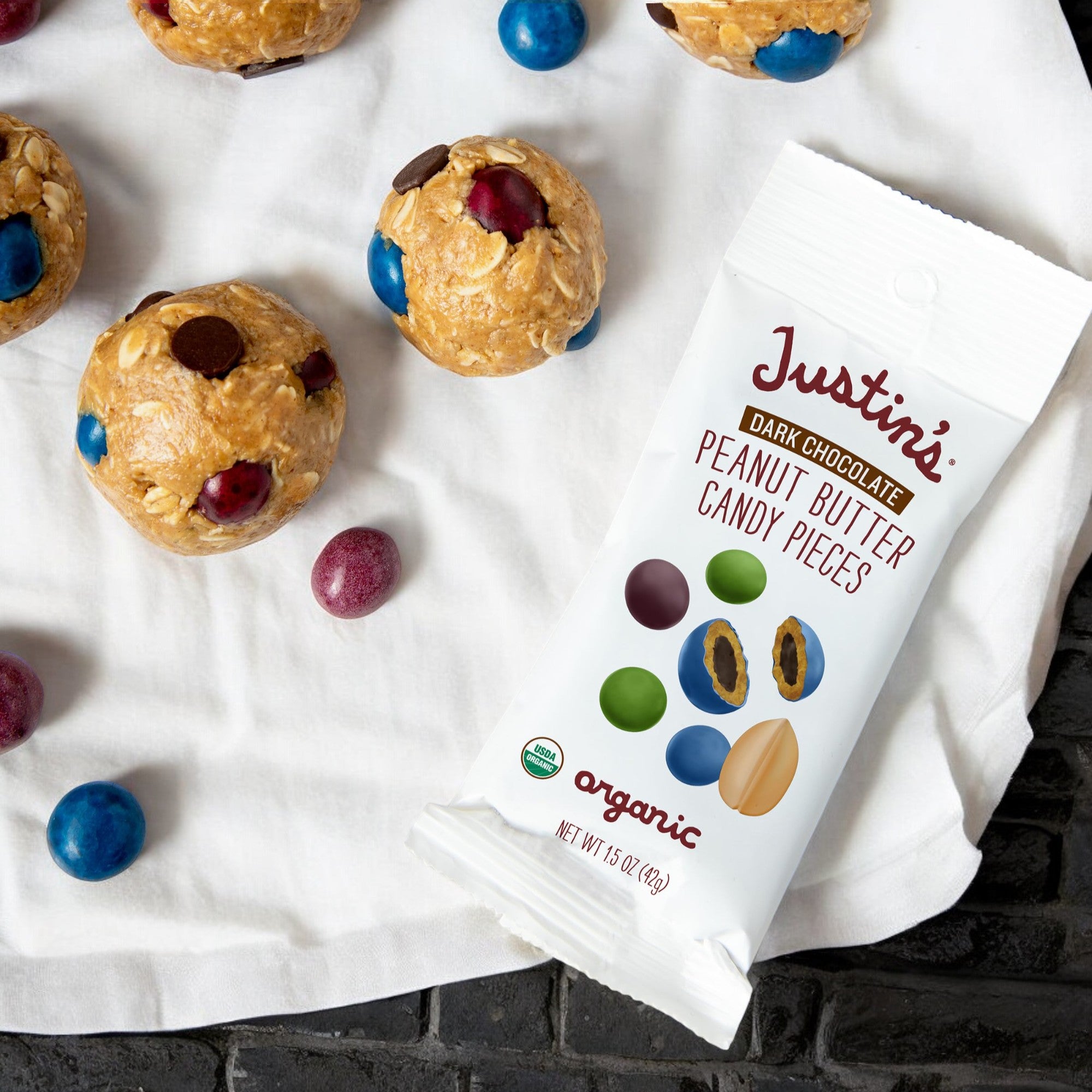 A package of Justin's Organic Peanut Butter Candy Pieces (1.5 oz, 1 count) from the reputable brand Justin's is showcased beside several spherical organic peanut butter snacks with candy pieces on a white cloth.