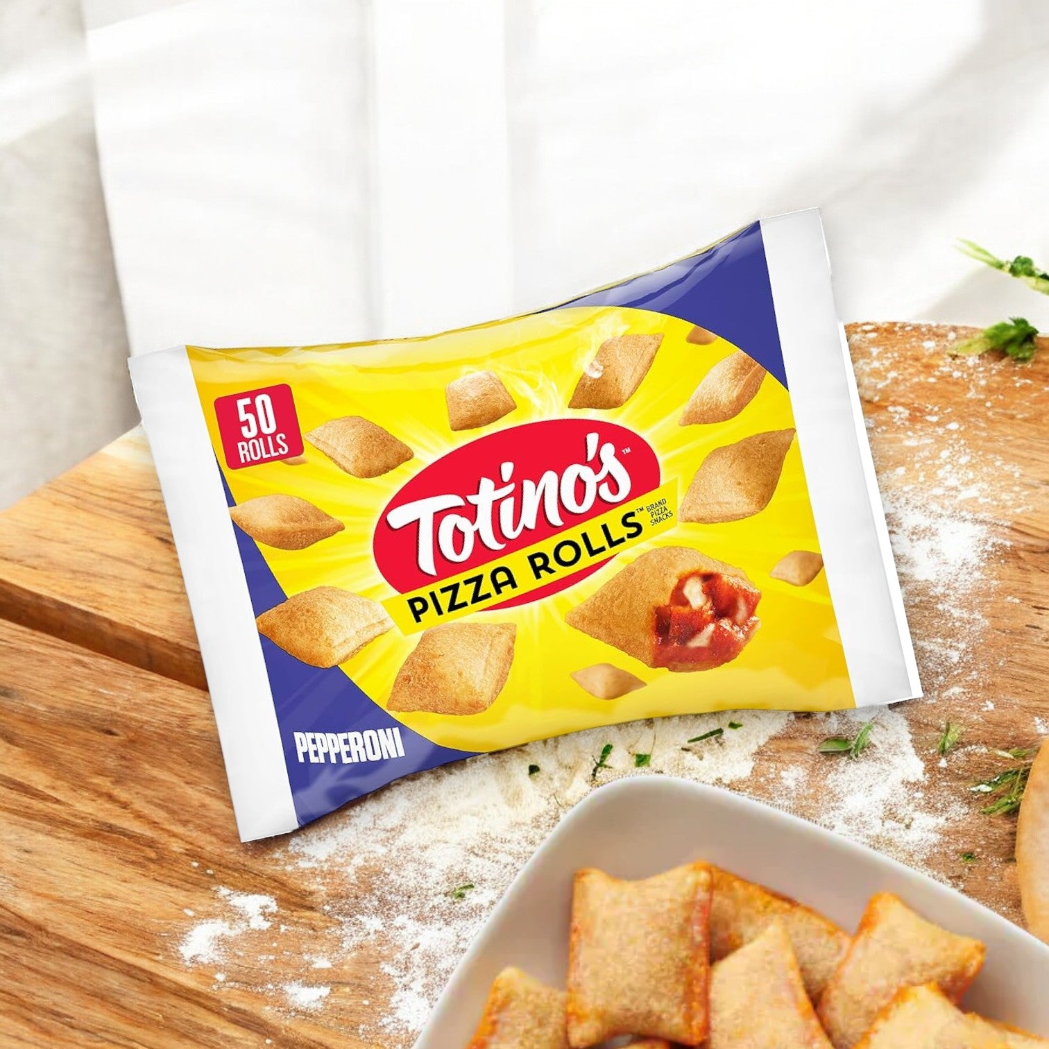 A package of Totino's Pizza Rolls, Pepperoni Flavored, Frozen Snacks, 24.8 oz, featuring 50 rolls, sits on a wooden cutting board. Nearby, some pizza rolls rest on a plate. A floured surface and herbs add to the classic pizza flavors ambiance in the background. Enjoy these Totino’s frozen snacks anytime!