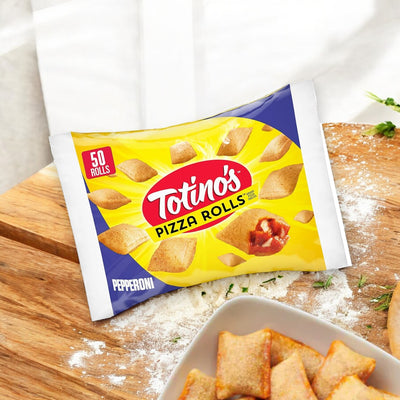 A package of Totino's Pizza Rolls, Pepperoni Flavored, Frozen Snacks, 24.8 oz, featuring 50 rolls, sits on a wooden cutting board. Nearby, some pizza rolls rest on a plate. A floured surface and herbs add to the classic pizza flavors ambiance in the background. Enjoy these Totino’s frozen snacks anytime!