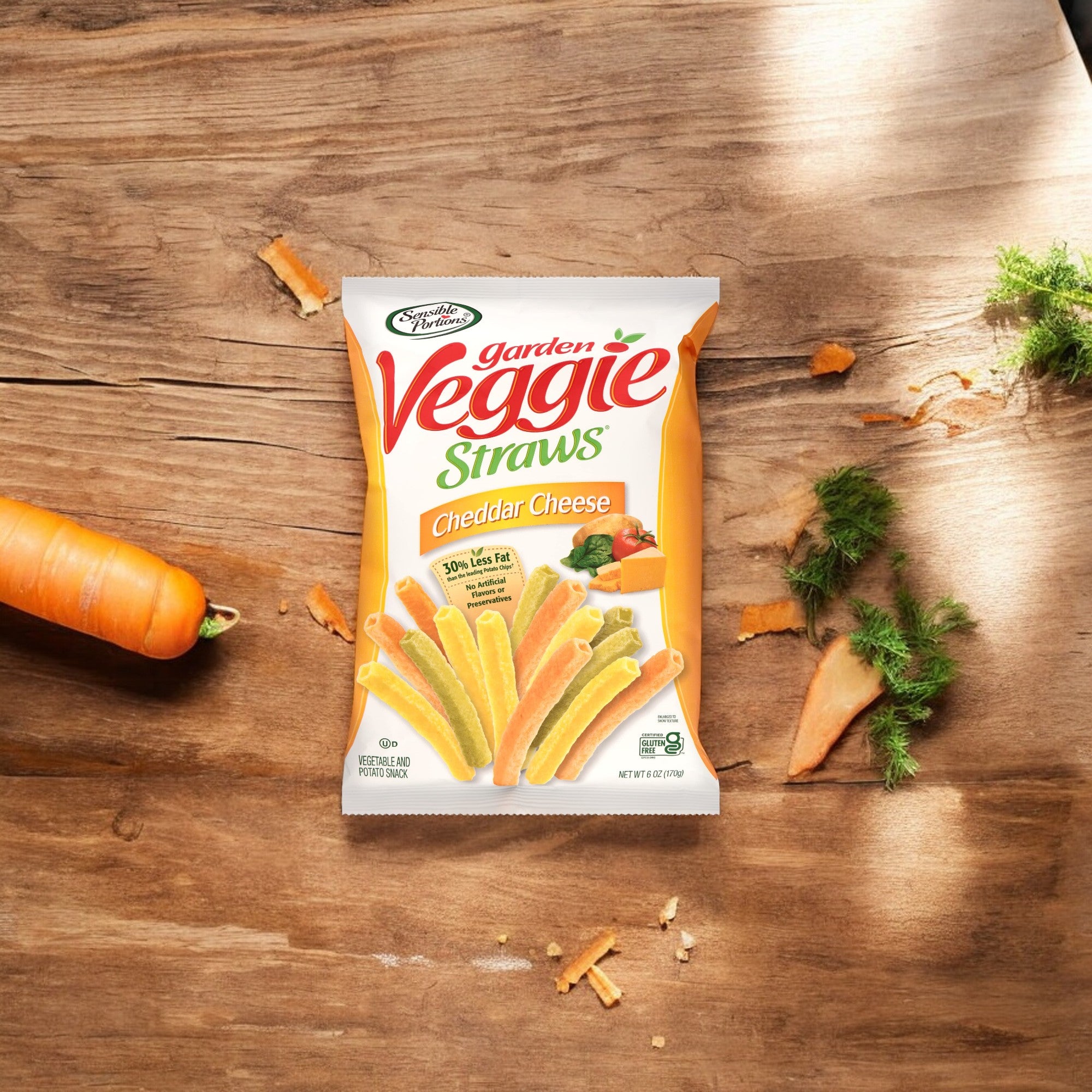 A bag of Hain Celestial's Garden Veggie Straws Cheddar Cheese (1 oz, 1 count) sits on a wooden surface surrounded by a partially peeled carrot and scattered carrot shavings.