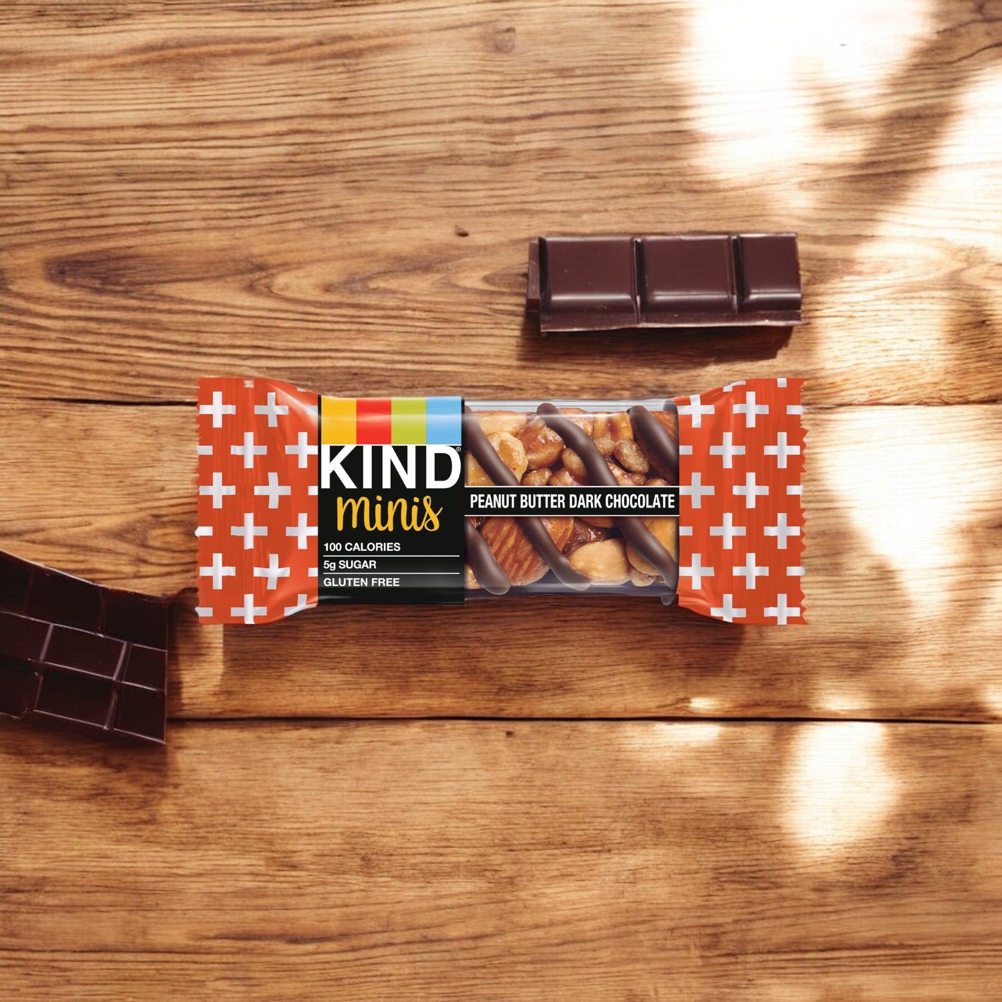 A Kind Minis Peanut Butter Dark Chocolate - 10 Count bar rests on a wooden surface, surrounded by scattered pieces of dark chocolate, making it an irresistible healthy snack.