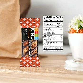 An image of a KIND Minis Peanut Butter Dark Chocolate 10 Count bar with a visible nutrition facts label displaying 100 calories per serving. The mini bar is set on a wooden surface beside a white vase with flowers, highlighting it as an ideal healthy snack.