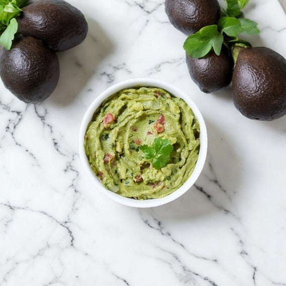 A bowl of Good Foods Chunky Guacamole - 1/2 oz garnished with a cilantro leaf, placed on a marble surface with four whole avocados arranged around it, makes for a delicious and healthy snack that's naturally gluten-free.