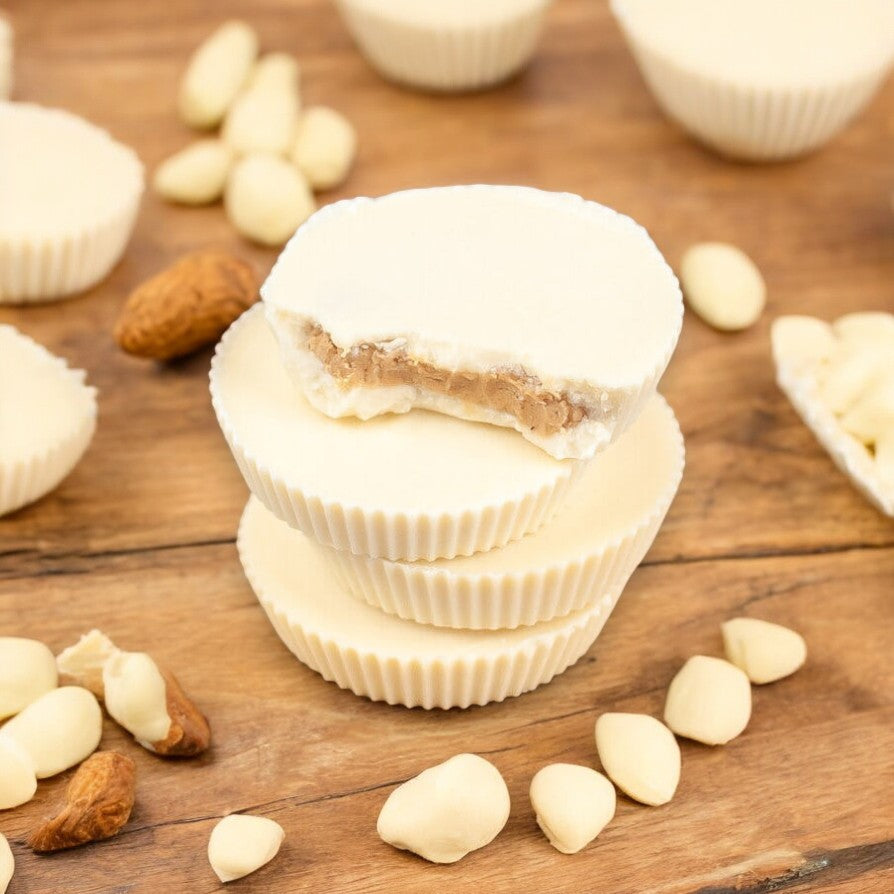 Three of Justin's Organic White Chocolate Peanut Butter Cups are stacked on a wooden surface, with one showing a bite taken out. Loose almonds and white chocolates scatter around, emphasizing the organic ingredients that deliver a pure, delightful taste.