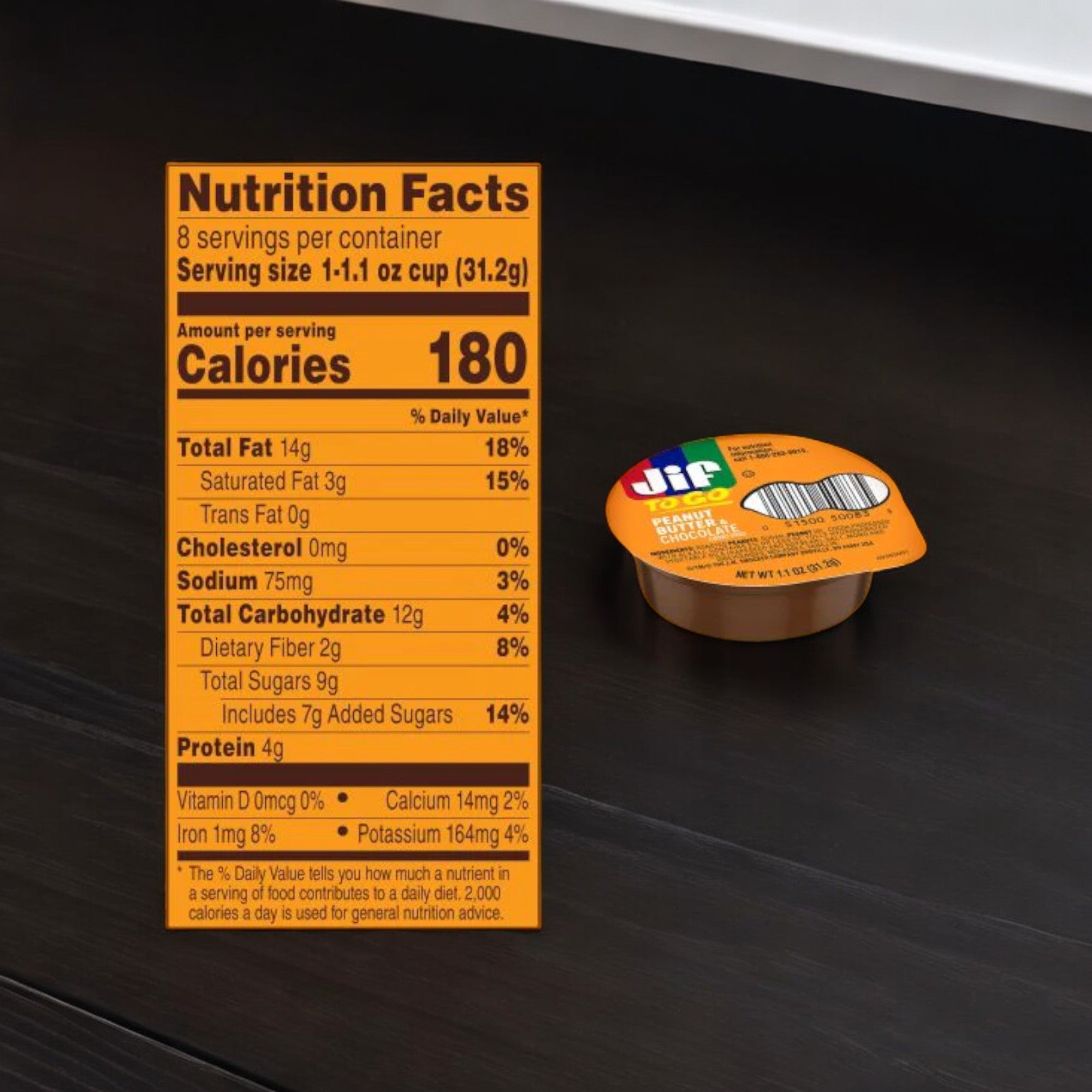 Photograph of a small container of Jif To Go Spread, Peanut Butter & Chocolate Flavored with the nutrition facts panel highlighted in the foreground. The panel indicates a serving size of 31.2g and 180 calories per serving, making it a tasty and healthy snack option compared to other chocolate-flavored spreads.