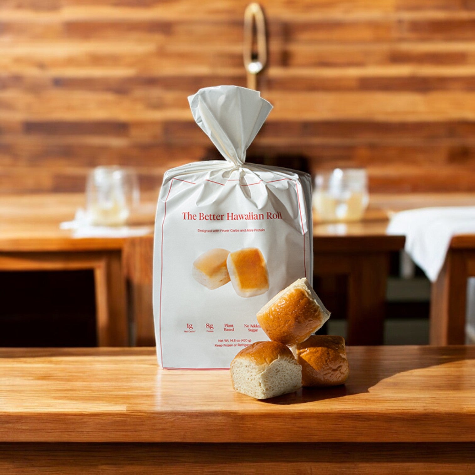 A pack of BetterBrand's "The Better Hawaiian Roll" made with premium ingredients is placed on a wooden table, accompanied by four stacked rolls. In the background, a wooden wall and blurred tableware highlight the signature sweetness found in each bite.