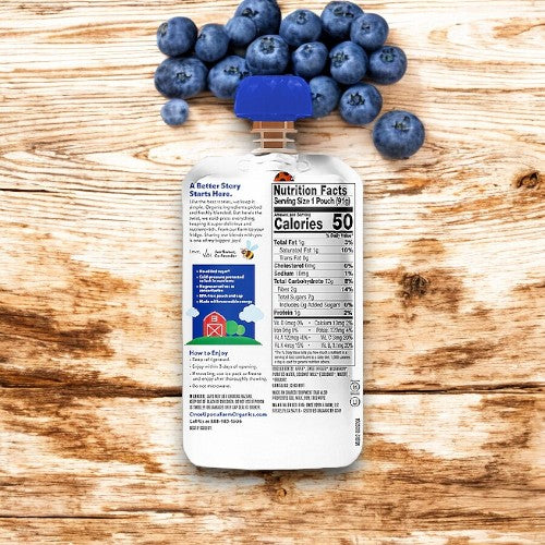 A stand-up pouch of Once Upon a Farm- Mama Blueberry baby food, featuring a blue cap, sits on a wooden surface. Topped with farm-fresh blueberries, the packaging emphasizes wholesome nutrition and includes nutritional information, noting a serving size of 1 pouch (99g) and 50 calories.