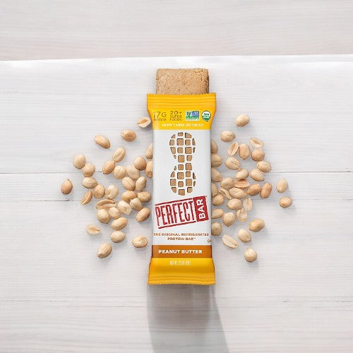 The Perfect Bar Gluten-Free Peanut Butter Refrigerated Protein Bar, 2.3 oz - 1 Count, in a yellow wrapper rests on a white surface, surrounded by scattered peanuts.