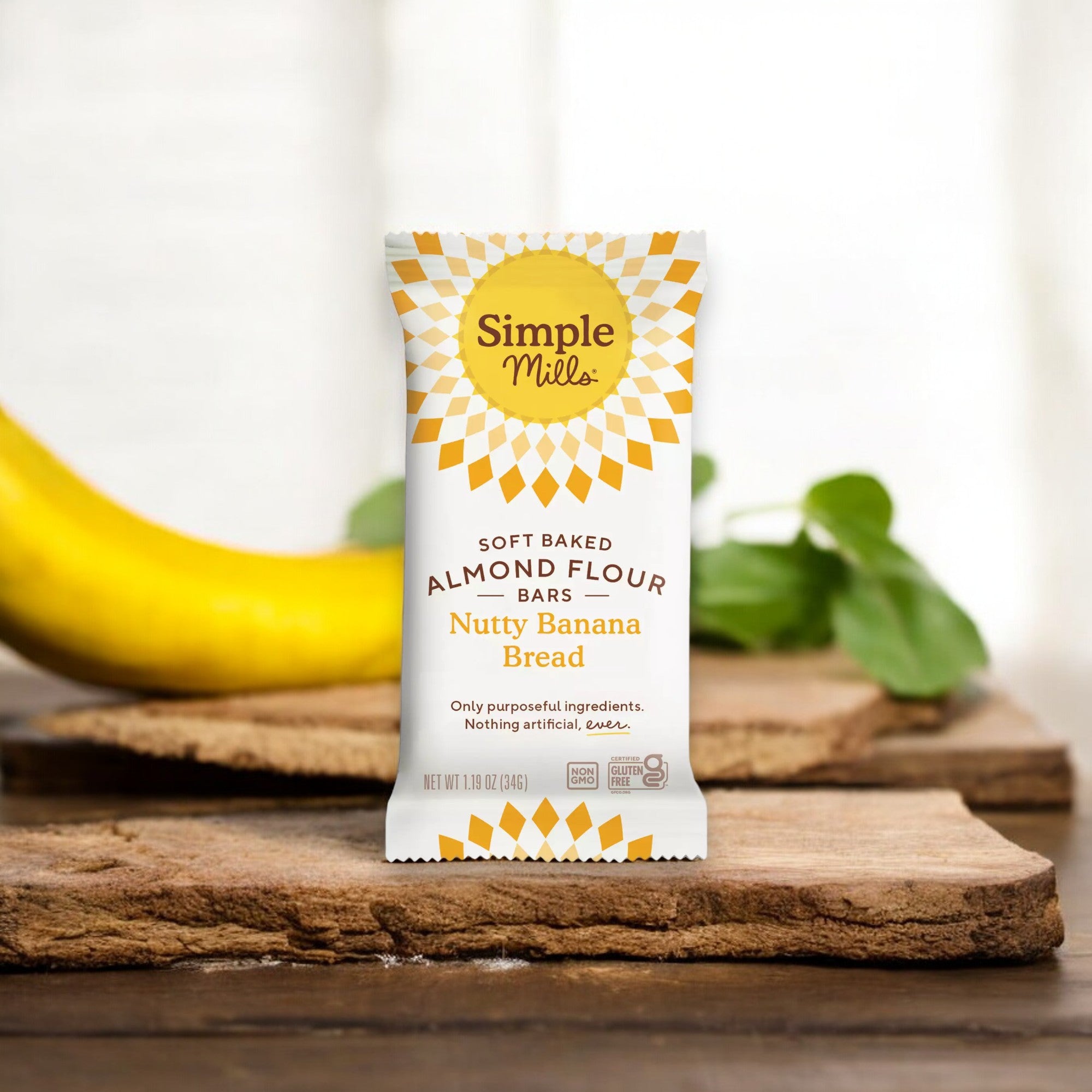 A packaged Simple Mills Soft Baked Almond Flour Bar, labeled "Nutty Banana Bread," is placed on a wooden board with a banana and green leaves in the background.