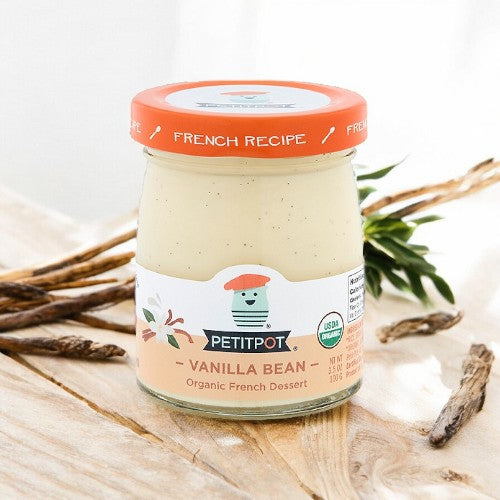 A jar of Petit Pot Organic French Vanilla Pudding sits on a wooden table beside vanilla beans and a green plant. The jar, with its orange lid and label adorned with text and illustrations, boasts the rich French Vanilla flavor derived from organic ingredients.