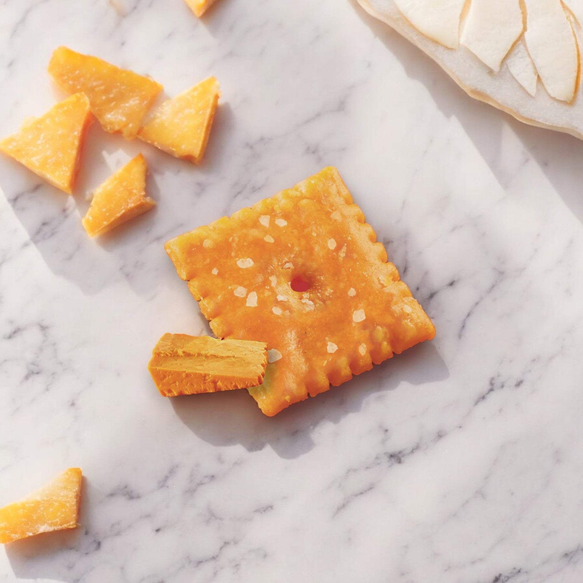 A Kellogg's Cheez-It Cheddar cracker and several pieces of real cheese are scattered on a white marble surface. A partially visible slice of white bread is in the upper right corner, promising a delightful Kosher snack.