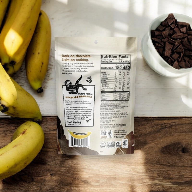 A bag of Barnana Banana Bites Organic Chewy Chocolate, 3.5 oz - 1 Count, is displayed on a wooden surface beside fresh bananas and a bowl of chocolate pieces. The back of the bag showcases product information and nutrition facts, emphasizing it as a sustainable snack option from the Barnara brand.